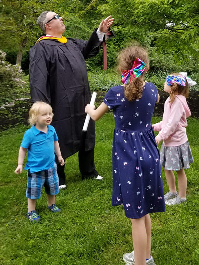 Larry Flagler, in a Commencement robe, tands outside with his family.