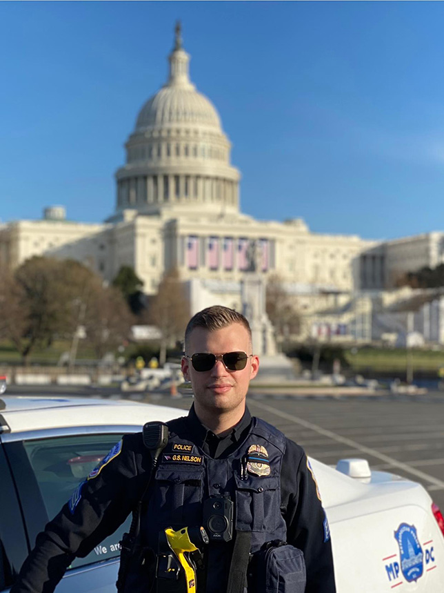 Gavin Nelson, in a police uniform, stands in front of the U.S. Capitol.