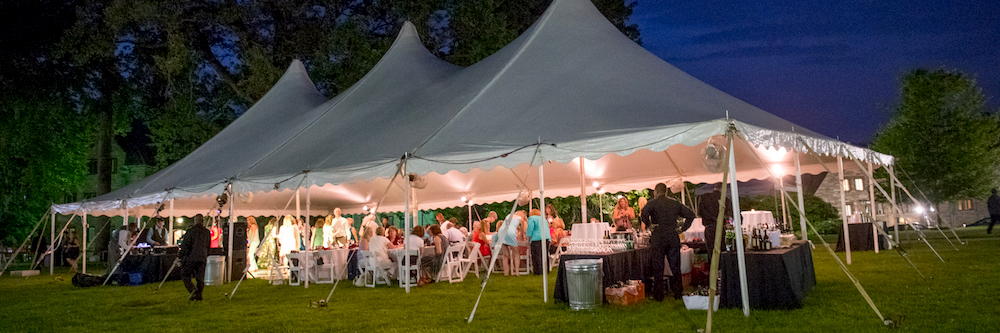 A white event tent, lit up filled with people attending a formal party/event. 