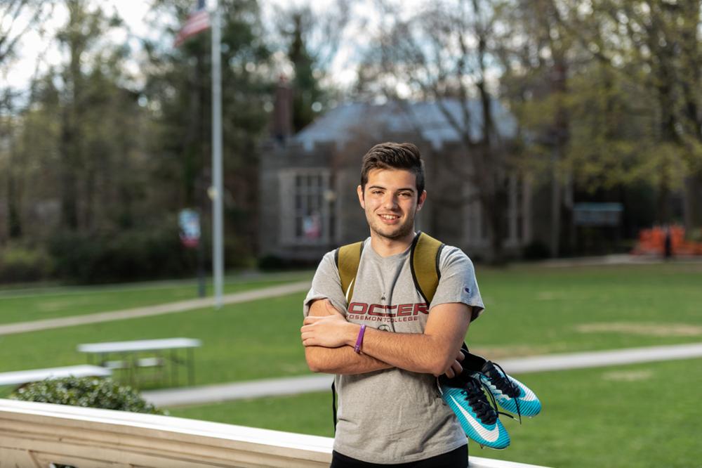 Zach, a Sport Management major at Rosemont has started a podcast of his own. He is pictured in the quad, and holds a pair of kleats. He is white and wears a grey t-shirt that says Rosemont on it. It is Spring or Summer.