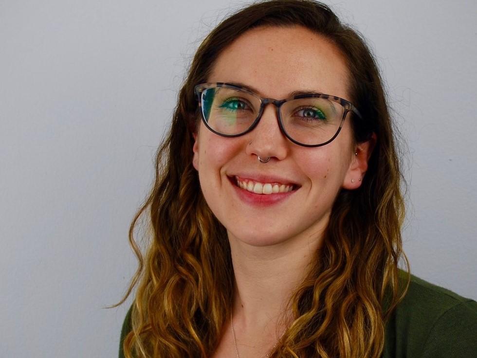 Image of Cassie Drumm, a young white woman with glasses and brown hair standing in front of a shelf of books.