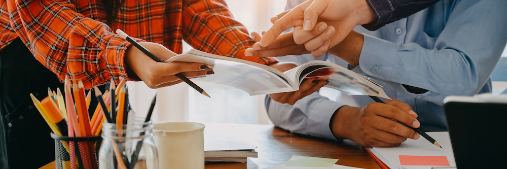 Image of three people's hands working together on a creative publishing project in an advertising office. There are pens and pencils, papers, and a computer on the desk. Two of the people are female, one is male with olive/tan skin.