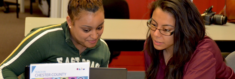 A young woman with medium brown skin and curly hair pulled back in a ponytail faces the direction of the camera. She looks down at some schoolwork on a desk. Another woman of latinx descent sits to her right, and leans toward her assisting her. She has mid-length straight black hair and glasses.