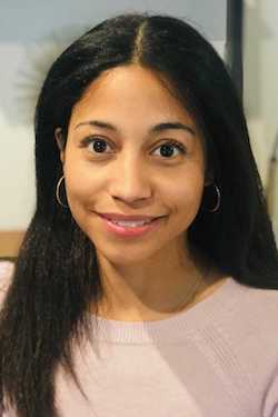 Portrait of Dr. Christina Clarke, a young woman of color with long black hair, light brown skin, and hoop earrings. She wears a light pink sweater,