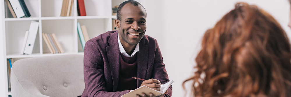 African American male therapist smiles at a patient across from his chair in a white, minimalist office. The back of the patient's head is visible, she is red-haired woman. A man's arm is slung around her shoulders. He has white skin.