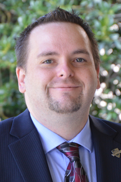 Image of Jay Kolick, a white man in his thirties with faint facial hair and short brown hair. He wears a dark blue suit and a tie with patterns on it.