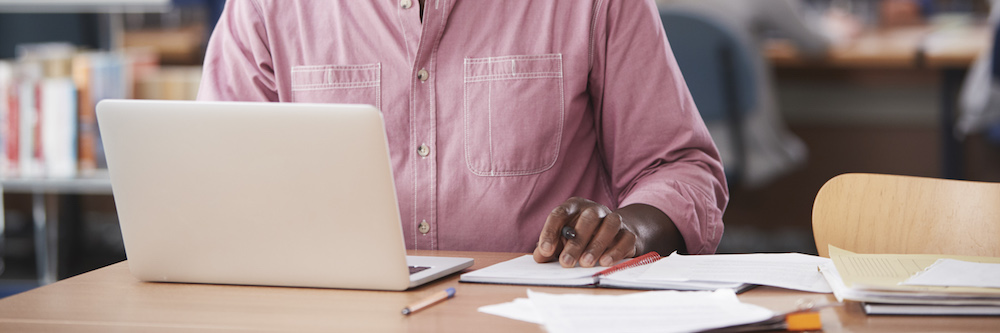 A pair of hands are pictured working on a laptop at a table. They belong to a an African-American male, and he wears a long-sleeved pink button down shirt.