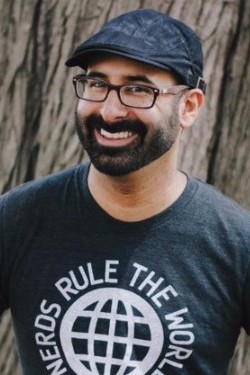 Eric Smith, Adjunct Faculty member in the MFA program at Rosemont. He is a tan/white man, wearing a hat, dark-rimmed glasses, beard, and a t-shirt that reads "Nerds Rule the World".