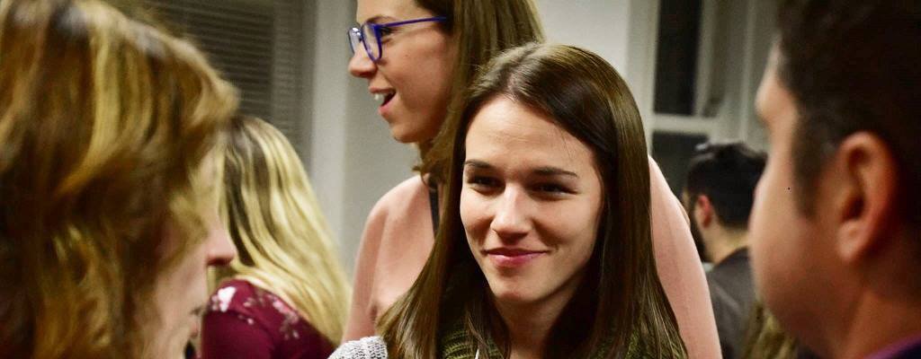 Image of a social gathering. Centered in the picture of a twenty-something brunette white woman. 