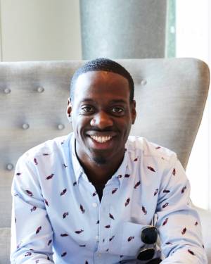 Leahcim James. He is a black man with short hair, wearing.a white shirt. He is sitting on a gray chair indoors. 