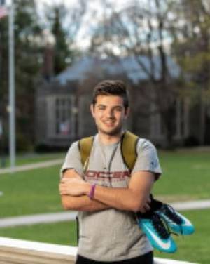 zach Young, a white male with short brunette hair, stands on a field. He's holding a pair of soccer shoes.