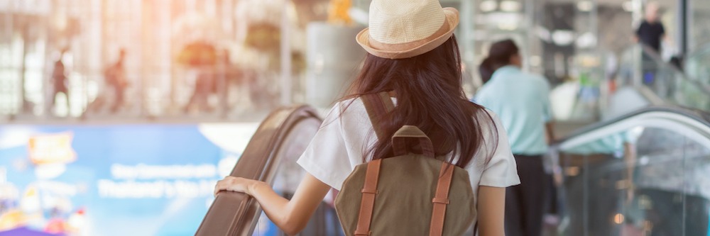 Young woman is pictured from behind as she ascends an elevator in the airport. She wears a light green t-shirt, a woven wide-brim hat, and a canvas backpack,