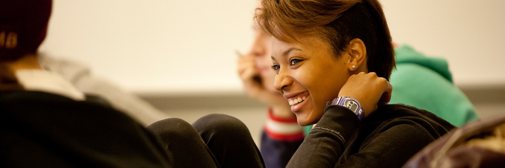 African-American woman leans forward on her desk in a college classroom. She has reddish highlights in her short hair and appears to be in her late-twenties.