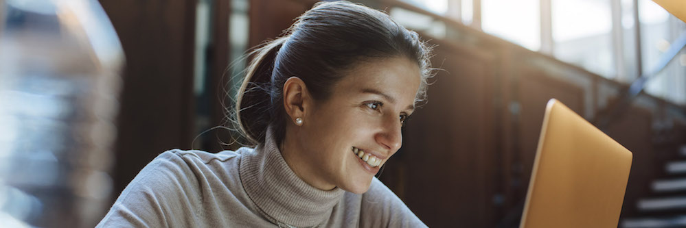 A young woman with light brown hair sits at a laptop in an academic setting. She wears a small cross necklace and a long-sleeved light pink shirt.