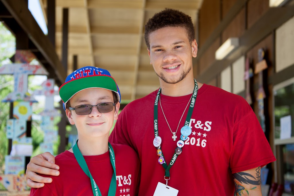 Photo of college-aged male with his arm around a younger boy who wears a red hat and glasses. Both wear red shirts and lanyards, and appear to be at a service event.